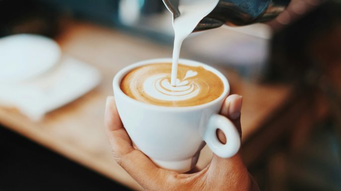 En hand häller mjölk i en kopp kaffe med en konstnärlig latteart-design.